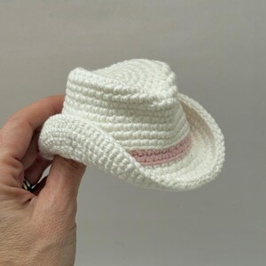 Peut inclure: Un petit chapeau de cowboy crochet&eacute; en blanc, pr&eacute;sentant un motif textur&eacute; et cousu sur toute la calotte et le bord. Une fine bande rose clair est int&eacute;gr&eacute;e autour de la base de la calotte, offrant un contraste de couleur subtil au fil blanc.
