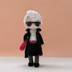 May include: Crocheted doll of a fashion icon, wearing black sunglasses, a black coat, and a red handbag. The doll has white hair and is wearing a white shirt, black skirt, and gray leggings. The doll is approximately 6 inches tall.