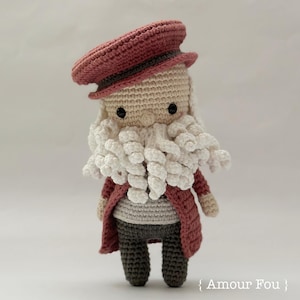 May include: A handmade crochet doll of a bearded man wearing a red hat and coat. The doll has a white beard, black eyes, and is dressed in gray pants and a gray shirt. The doll is approximately 6 inches tall.