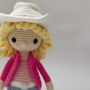 Peut inclure: Une poup&eacute;e crochet&eacute;e &agrave; la main avec un chapeau de cowboy cr&egrave;me, des cheveux en laine jaune boucl&eacute;s et deux yeux en perles noires. La poup&eacute;e porte une veste rose vif sur un haut rose clair et un pantalon bleu, avec une boucle de ceinture blanche.