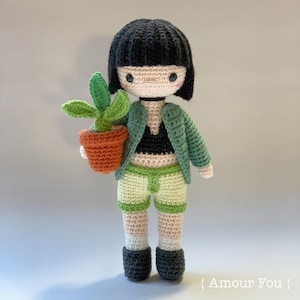 Puede incluir: Una muñeca de crochet con cabello negro, que lleva una chaqueta verde, un top negro y pantalones cortos verdes. La muñeca sostiene una pequeña planta en maceta con hojas verdes.