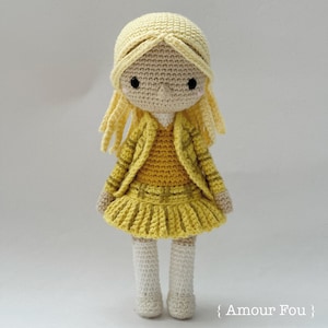 May include: A crocheted doll with blonde hair wearing a yellow dress and jacket. The doll has a white background.
