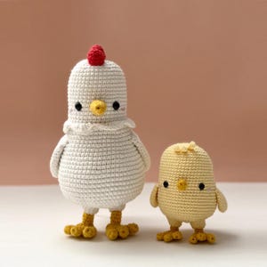 May include: Two crocheted chicken figures. One is white with a red comb, yellow beak, and feet. The other is yellow with a similar design. Both have black eyes and are standing upright.