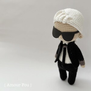 May include: A crocheted doll wearing a white hat, black sunglasses, and a black suit. The doll has a white shirt and a black tie. The doll is standing on a white surface.