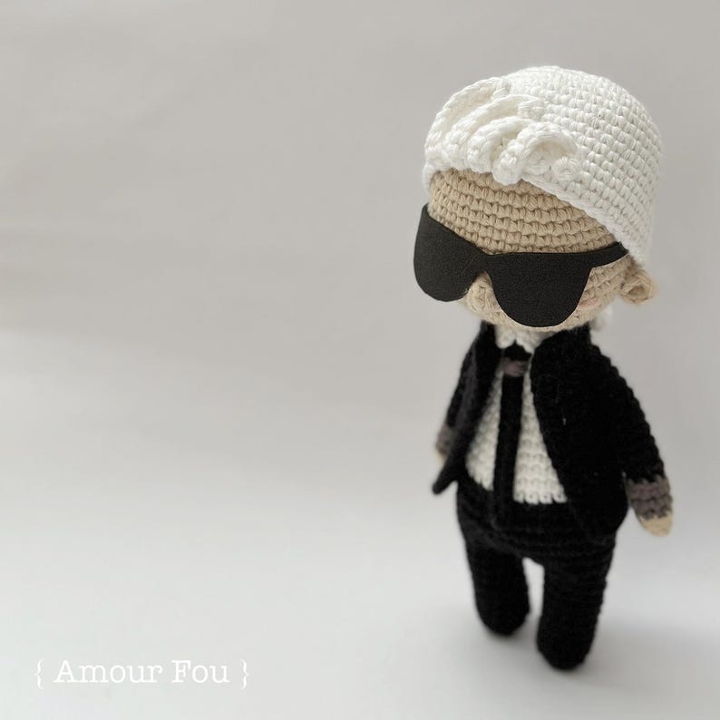 May include: A crocheted doll wearing a white hat, black sunglasses, and a black suit. The doll has a white shirt and a black tie. The doll is standing on a white surface.