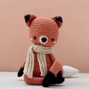 May include: A crocheted fox doll in shades of orange, black, and white, wearing a beige scarf. The fox has black eyes and ears, and a white-tipped tail. The doll is sitting down.