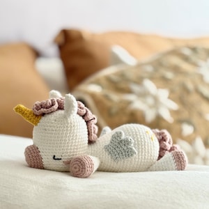 May include: A white crocheted unicorn with pink and yellow details, lying on a white bedspread. The unicorn has a pink mane, a yellow horn, and a light blue wing.