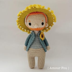May include: A crocheted doll with a yellow hat, orange hair, and a blue jacket. The doll is holding a small sunflower.