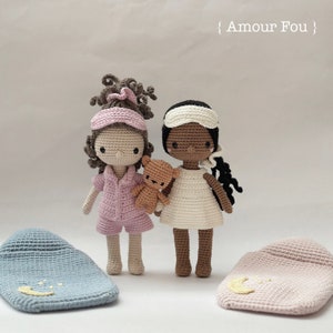 May include: Two crocheted dolls wearing pajamas and sleep masks, holding a teddy bear. The dolls are standing on a white surface with two crocheted sleeping bags.