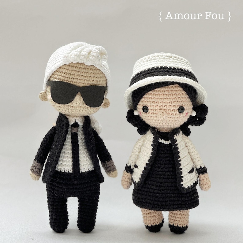May include: Two crocheted dolls, one wearing a black suit and sunglasses, the other wearing a white jacket and black dress. The dolls are standing on a white background. The text "Amour Fou" is written above the dolls.