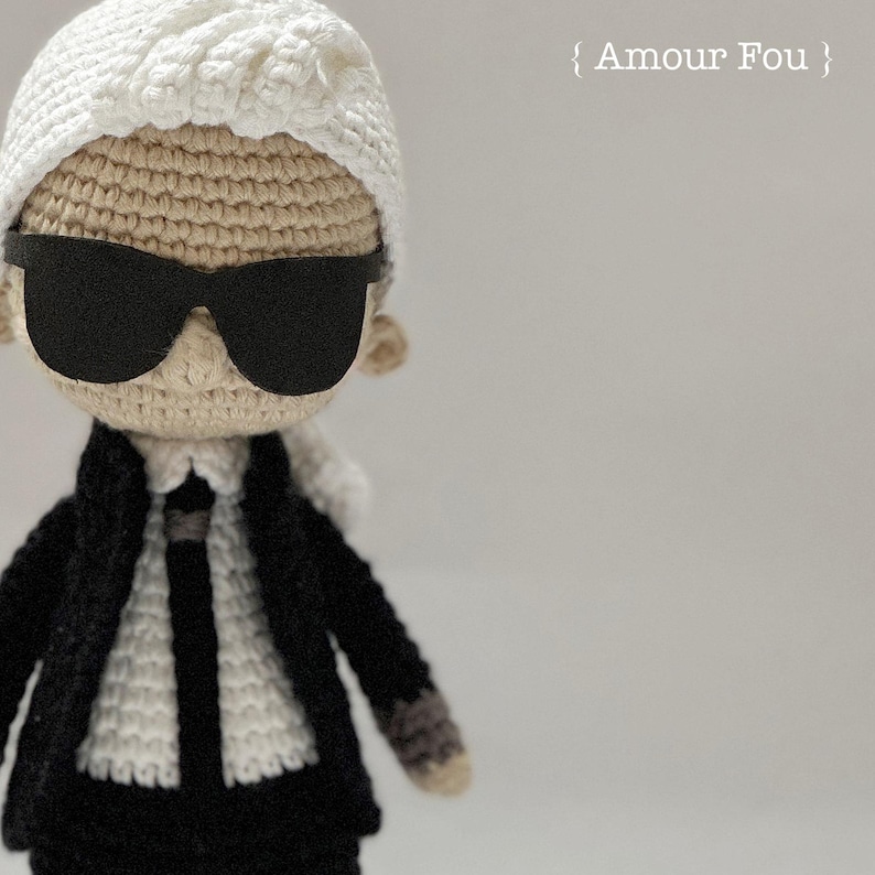 May include: A crocheted doll wearing a white hat, black sunglasses, and a black suit with a white shirt and black tie. The doll is standing on a white background. The text "Amour Fou" is written in white on a black background.