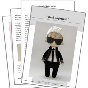 May include: A crochet amigurumi doll of Karl Lagerfeld, wearing a black suit, white shirt, black tie, and sunglasses. The doll is standing on a white background with the text "Karl Lagerfeld" above it.