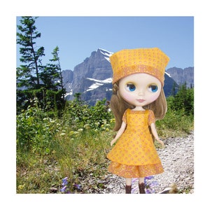 May include: A doll wearing a yellow and orange floral dress and a matching hat. The doll has blue eyes and is standing in a field with mountains in the background.