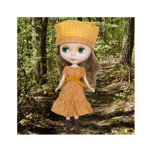 Puede incluir: Una muñeca con un vestido floral amarillo y naranja con una diadema amarilla a juego. La muñeca tiene el pelo largo y castaño y grandes ojos azules. La muñeca está de pie en un entorno de bosque.