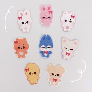 Könnte beinhalten: Eine Sammlung von acht Cartoon-Tieraufklebern. Die Aufkleber zeigen eine Vielzahl von Tieren, darunter ein Hase, ein Schwein, eine Katze, ein Bär und ein Hund, jedes mit einem niedlichen Design und einer kleinen Schleife. Die Farbpalette umfasst Rosa, Orange, Gelb, Blau und Beige.