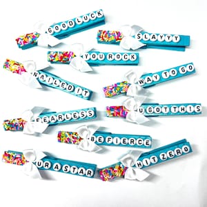 May include: Ten turquoise clothespins with white bows and colorful sprinkles. Each clothespin has a different motivational message in black letters, such as "Good Luck", "You Rock", "Way To Go", and "Be Fierce".