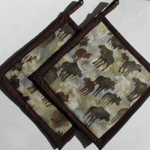 May include: Two brown and beige pot holders with a moose and other animal silhouette pattern. The pot holders have a dark brown border.