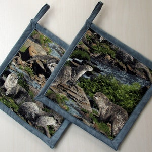 May include: Two blue and gray fabric pot holders with a nature scene featuring otters and a stream. The fabric has a textured look.