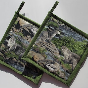 May include: Two green fabric pot holders with a nature scene featuring otters and rocks. The pot holders have a loop for hanging.