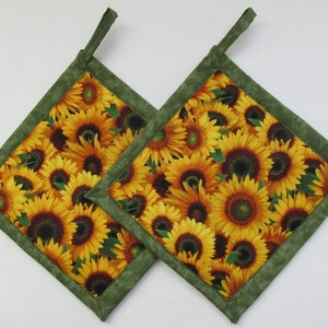 May include: Two square pot holders with a sunflower pattern. The pot holders have a yellow and brown sunflower design with a green border and a green loop for hanging.