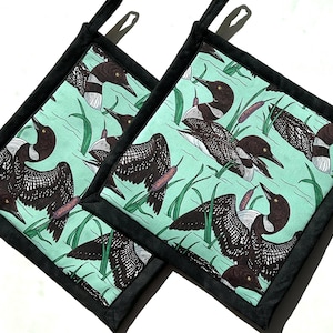 May include: Two square pot holders with a teal background featuring a pattern of black and white loons, cattails, and green reeds. The pot holders have a black border and a black hanging loop.