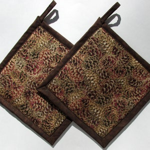 May include: Two square pot holders with a floral pattern in shades of brown, beige, and burgundy. The edges are trimmed with a solid brown fabric, and each has a small loop for hanging. Kitchen and dining decor.