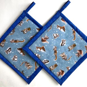 May include: Two square pot holders with a light blue background featuring a pattern of squirrels and snowflakes. The pot holders have a dark blue border and a loop for hanging. The pot holders are made of fabric.
