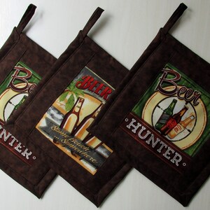May include: Set of three brown fabric pot holders with a green, yellow, and white design featuring beer bottles and the text "Beer Hunter".