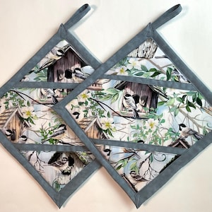 May include: Two diamond-shaped pot holders featuring a pattern of small birds perched on wooden birdhouses amidst green leaves and white flowers. Each pot holder has a solid gray fabric border and a matching gray fabric hanging loop at the top.