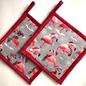 May include: Two square potholders with a red border and hanging loop. The gray fabric features pink flamingos wearing winter hats and scarves, with snowflakes. The potholders are designed for kitchen use.