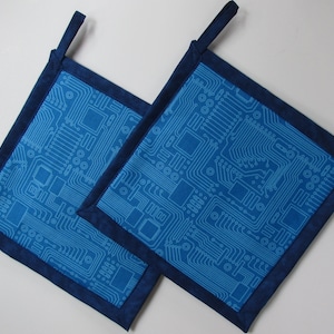 May include: Two square pot holders with a blue circuit board pattern on a lighter blue background. The edges are trimmed with a darker blue fabric, and each has a small loop for hanging.