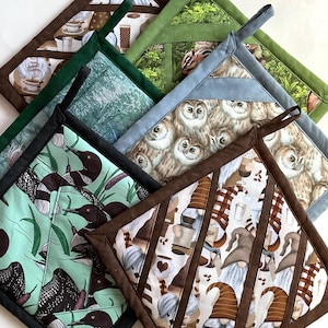 May include: A collection of square pot holders with various patterns. Designs include owls, gnomes, loons, and coffee cups. Each pot holder has a contrasting border and a loop for hanging. The color palette includes greens, blues, browns, and white.