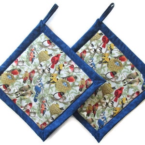 May include: A pair of square pot holders featuring a vibrant bird and nest design on a light backdrop, edged with a dark blue border. Each has a dark blue loop for hanging.