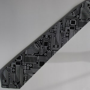 May include: Black necktie with a white circuit board pattern. The tie features a repeating design of electronic components and pathways, creating a tech-inspired aesthetic. The tie is angled across a white background.