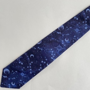 May include: A dark blue tie featuring a celestial design of white stars and crescent moons. The tie is angled diagonally against a white backdrop. The fabric has a subtle texture, adding depth to the design.