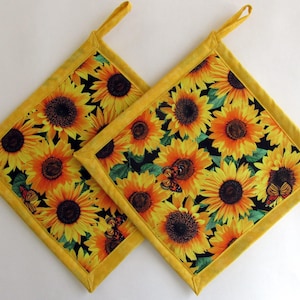 May include: Two square pot holders with a vibrant sunflower pattern. The design features yellow sunflowers with orange centers and green leaves against a black background. Each pot holder has a yellow border and a loop for hanging.