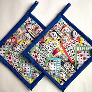 May include: Two square pot holders with a blue border feature a colorful bingo-themed fabric. The fabric displays bingo cards and bingo balls. Each pot holder has a blue loop for hanging.