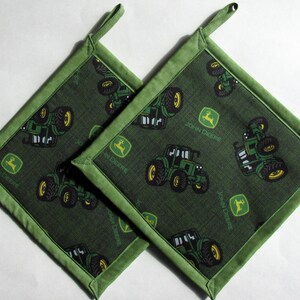 May include: Two square green pot holders with a John Deere tractor pattern. The pot holders have a dark green background with green tractors and yellow wheels. The edges are trimmed with a lighter green fabric and have a loop for hanging.
