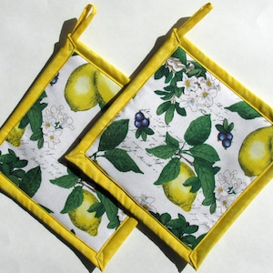May include: Two square pot holders with a white background featuring lemons, green leaves, and white flowers. The pot holders have a yellow border and a yellow hanging loop. Kitchen decor.