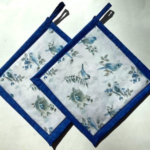 May include: Two square pot holders featuring a blue bird and berry pattern on a white backdrop. They have a dark blue border and hanging loop. The design includes birds, nests, and berries.