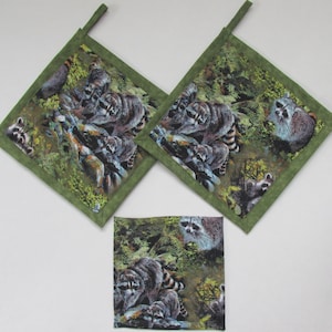 May include: Set of three green and brown pot holders with a raccoon print. The pot holders have a green border and a hanging loop.