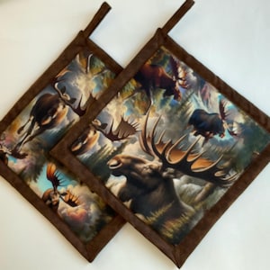 May include: Two square pot holders with a brown border and hanging loop. The center features a detailed image of moose in a forest setting. The pot holders are made of fabric and are designed for kitchen use.
