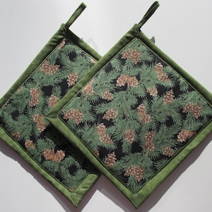 May include: Two green and black quilted pot holders with a pine cone and evergreen print. The pot holders have a green border and a loop for hanging.