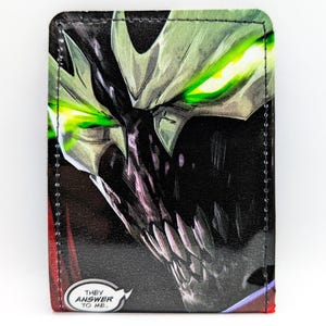 May include: A rectangular wallet featuring a graphic illustration of a dark, masked character with glowing green eyes and sharp teeth. The design includes a speech bubble in the bottom corner with the text, "THEY ANSWER TO ME."