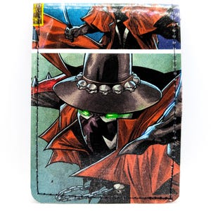 May include: A rectangular wallet featuring a comic book illustration of a character wearing a wide-brimmed hat adorned with skulls, a red cape, and glowing green eyes. The design includes a chain detail and black stitching along the edges.