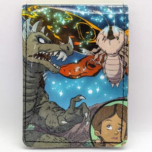 May include: A blue and green wallet with a cartoon image of a monster, a butterfly, and a human character. The monster has a large mouth with sharp teeth and is green and gray. The butterfly is white and has a brown body. The human character is brown-skinned and has brown hair.