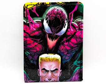 May include: A rectangular wallet featuring a comic book illustration of a character with a white face, sharp teeth, and a pink and black design. Below is a man with blonde hair and blue eyes. The background is black.