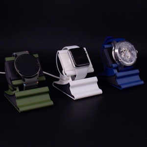 Peut inclure: Trois supports de montre, chacun tenant une montre intelligente différente. Les supports sont verts, blancs et bleus. Les montres ont des cadrans noirs, blancs et argentés. Les supports sont sur une surface noire.