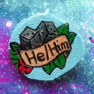 May include: A hand-painted round wooden pin with a blue background. The pin features a cluster of gray dice with green leaves and a banner that reads "Hell Him".