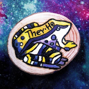 May include: A wooden circle with a painted frog on it. The frog is white with purple and yellow stripes and spots. The frog has the words "They/He" written on its back.